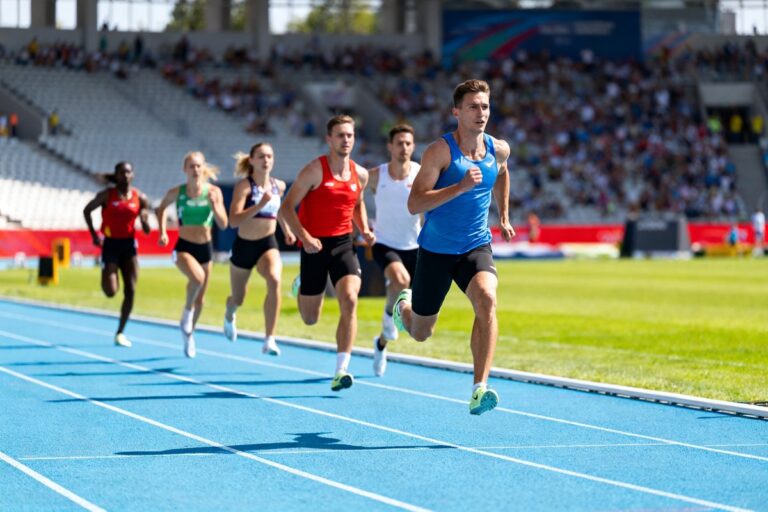 Six coureurs et coureuses lancés à pleine vitesse sur une piste d'athlétisme bleue devant des tribunes remplies