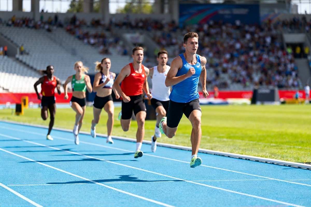 Six coureurs et coureuses lancés à pleine vitesse sur une piste d'athlétisme bleue devant des tribunes remplies