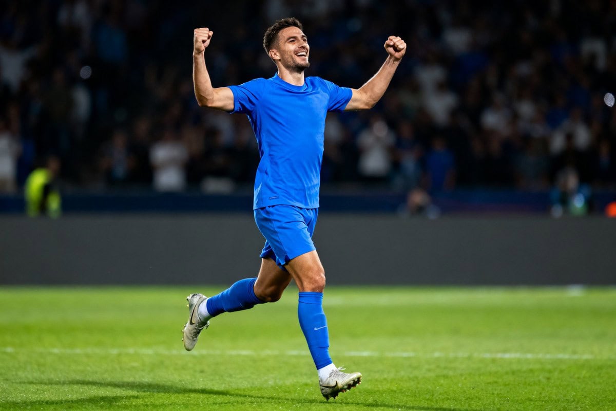 Un footballeur en maillot bleu uni, bras levés, célèbre un but en courant sur une pelouse de stade illuminée la nuit.