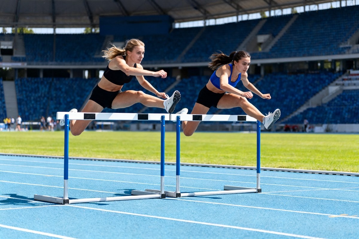 Deux hurdleuses franchissent une haie côte à côte sur une piste bleue, en plein vol