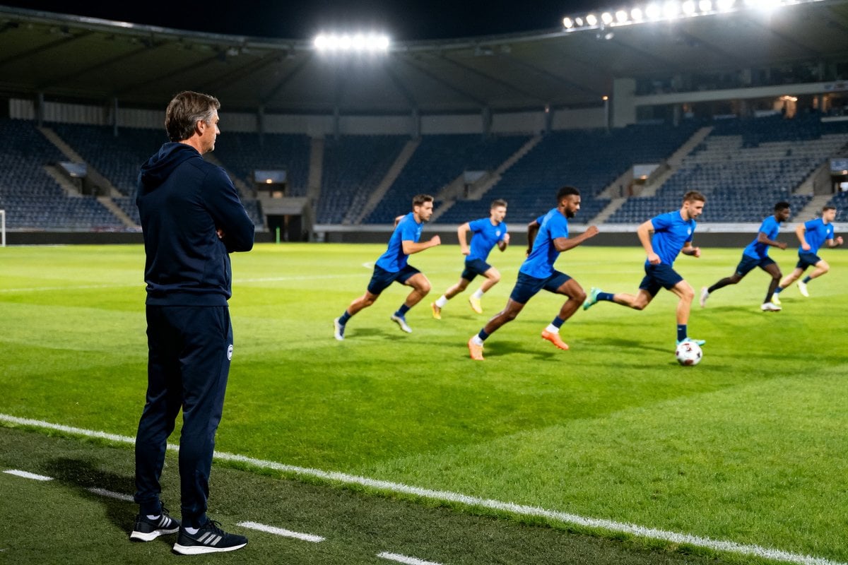 Des footballeurs en maillot bleu sprintent sur une pelouse éclairée, un entraîneur les observe depuis la touche