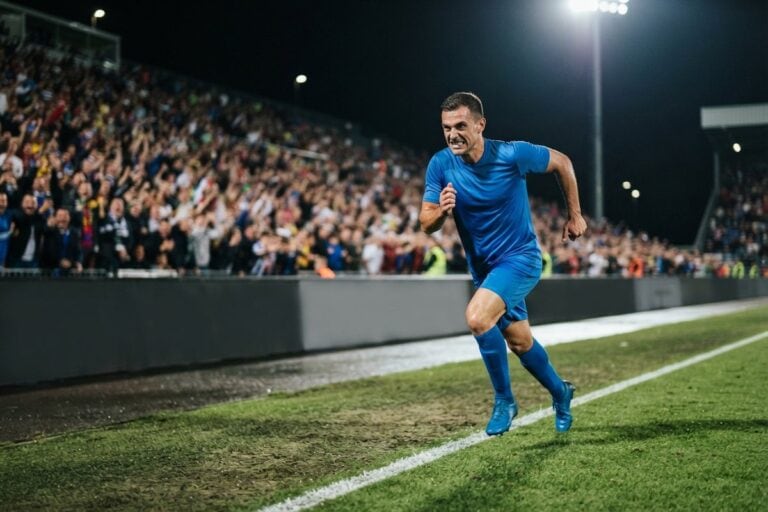 Un footballeur en tenue bleue court le long de la ligne de touche d'un stade éclairé, tribunes pleines derrière lui.