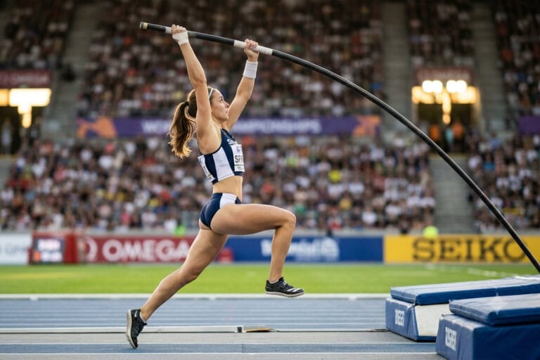 Une perchiste franchit la barre au-dessus d'un stade d'athlétisme rempli, sous un ciel d'été
