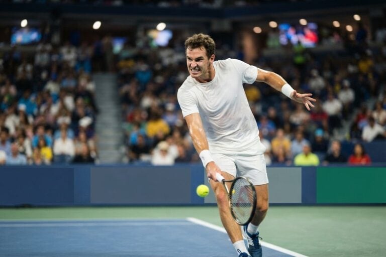 Un joueur de tennis frappe une balle basse sur un court en dur bleu, devant les tribunes d'un stade éclairé