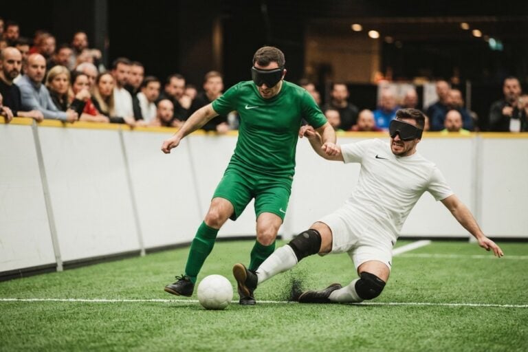 Deux joueurs de cécifoot masqués se disputent le ballon le long des balustrades d'un terrain couvert.