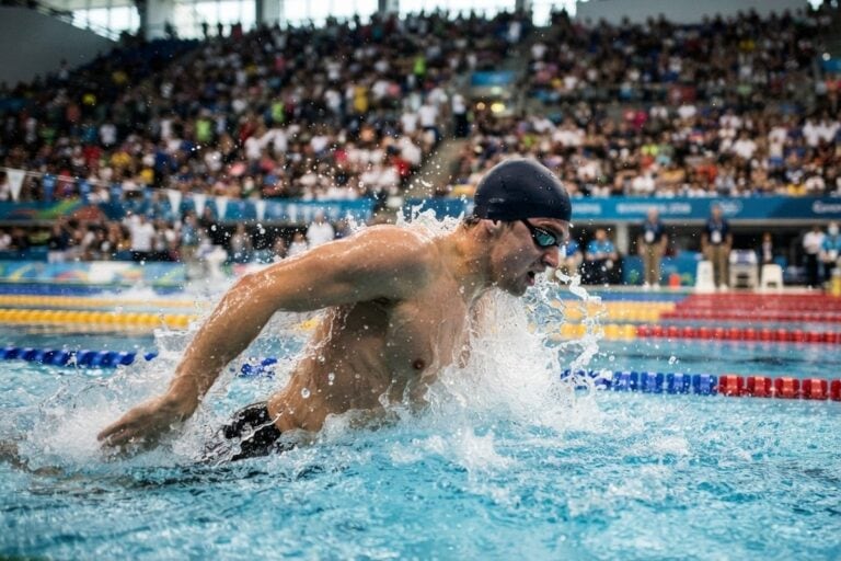 Un nageur en pleine action dans un bassin olympique, gerbes d'eau autour de lui, tribunes remplies