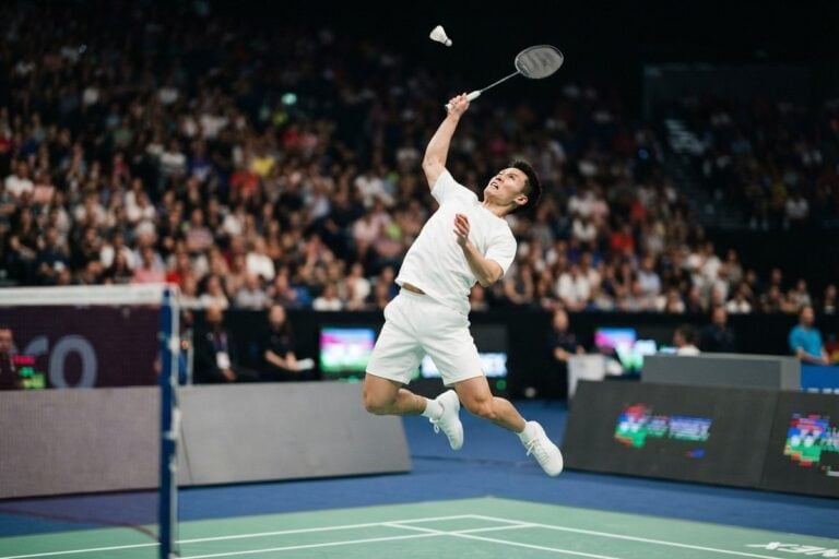 Un joueur de badminton en suspension au moment du smash, raquette au-dessus de la tête, devant un public dense
