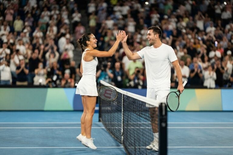 Une joueuse et un joueur de tennis en tenue blanche se tapent dans la main au-dessus du filet, devant un public debout.
