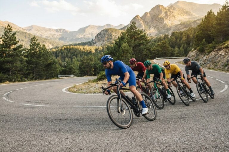 Cinq cyclistes en file dans un virage en épingle, sur une route de montagne bordée de pins et de sommets rocheux.