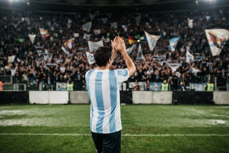 Un footballeur en maillot rayé bleu ciel et blanc, vu de dos, applaudit une tribune pleine de drapeaux
