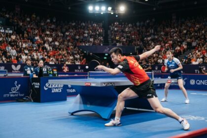 Un pongiste en maillot rouge frappe la balle sur une table bleue, devant des tribunes pleines
