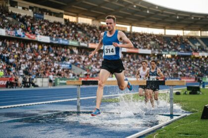 Coureur en maillot bleu retombant dans la rivière du steeple, deux concurrents derrière lui sur la piste