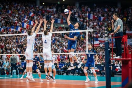 Un attaquant en maillot bleu frappe le ballon au-dessus d'un double contre en blanc, devant des tribunes pavoisées de drapeaux français