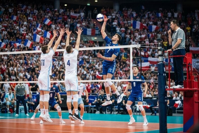 Un attaquant en maillot bleu frappe le ballon au-dessus d'un double contre en blanc, devant des tribunes pavoisées de drapeaux français
