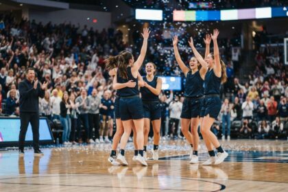 Cinq basketteuses en maillot bleu marine lèvent les bras au centre du parquet sous les confettis, devant un public debout