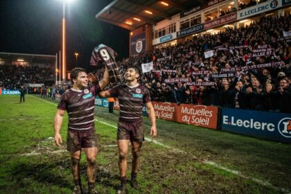 Deux joueurs de rugby en maillot bordeaux brandissent un maillot de match devant une tribune pleine