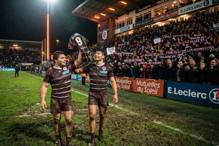 Deux joueurs de rugby en maillot bordeaux brandissent un maillot de match devant une tribune pleine