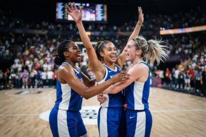 Trois basketteuses en maillot bleu et blanc s'étreignent sur le parquet après leur victoire, devant un public flou
