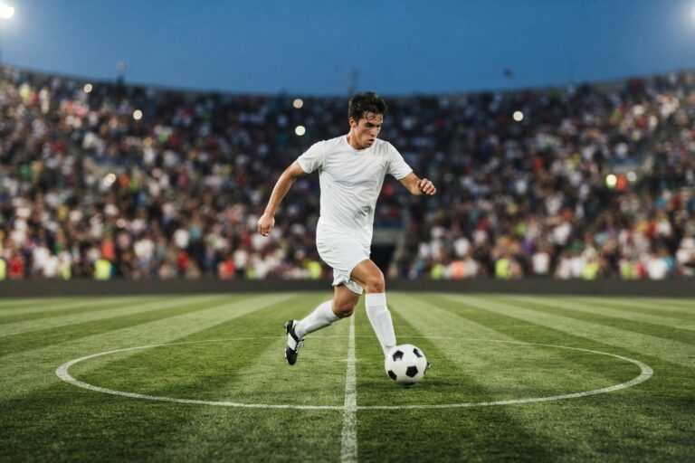 Un footballeur en maillot blanc conduit le ballon au rond central d'un stade éclairé, tribunes pleines derrière lui