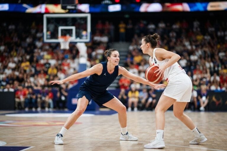 Une basketteuse en maillot bleu marine défend bras écartés face à une joueuse en blanc qui tient le ballon