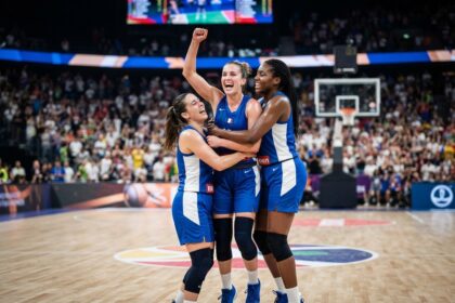Trois basketteuses en maillot bleu s'enlacent au centre du terrain, poing levé, devant des tribunes pleines