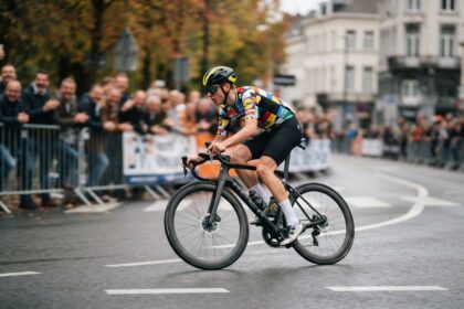 Un cycliste en maillot multicolore roule seul sur une route urbaine mouillée, devant des spectateurs derrière des barrières