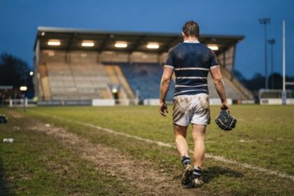 Un rugbyman en maillot rayé bleu marine quitte de dos un terrain boueux au crépuscule, casque de mêlée à la main