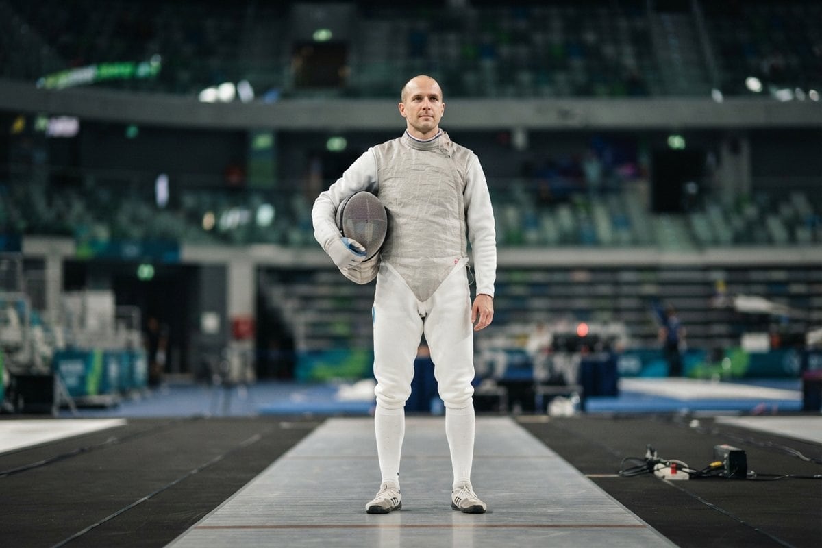 Un escrimeur en tenue blanche et veste lamée tient son masque sous le bras, debout sur une piste de salle