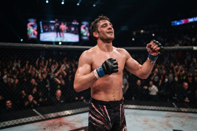 Combattant de MMA torse nu et gants ouverts, poings serrés au centre d'une cage, devant un public en tribunes