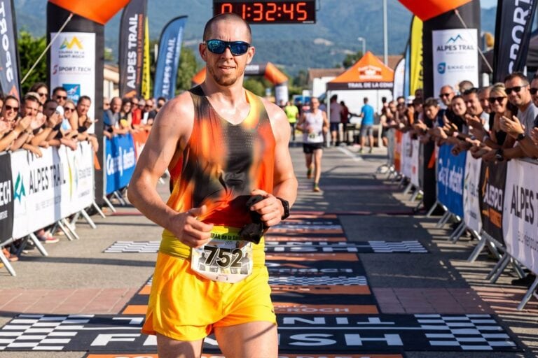 Un coureur en débardeur orange et short jaune franchit la ligne d'arrivée d'un trail sous les applaudissements