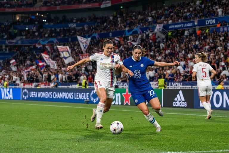 Une joueuse en maillot blanc de Lyon et une joueuse en bleu de Chelsea à la lutte pour le ballon, tribunes pleines