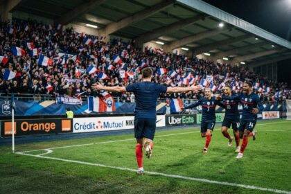Joueur de l'équipe de France de dos, bras écartés devant une tribune pleine de drapeaux tricolores, rejoint par trois coéquipiers