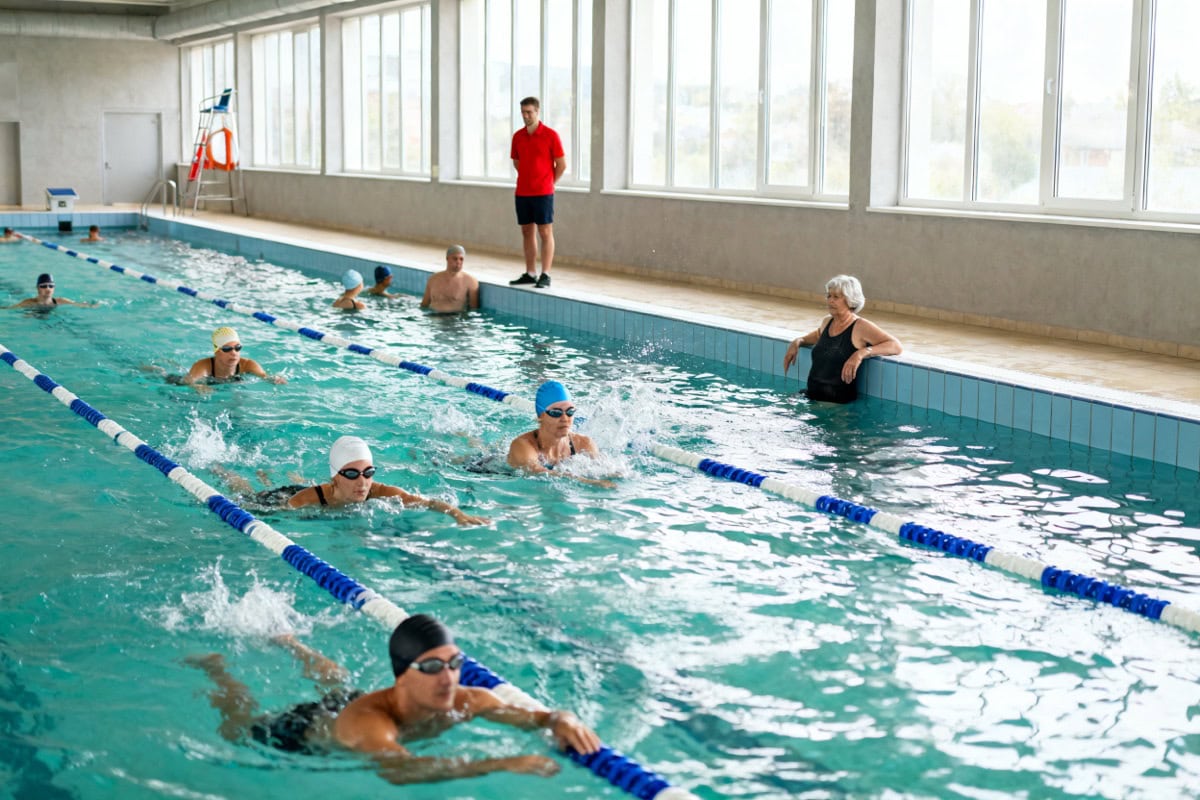 Nageuses et nageurs adultes faisant des longueurs dans le bassin de 25 mètres d’une piscine municipale, sous le regard d’un maître-nageur