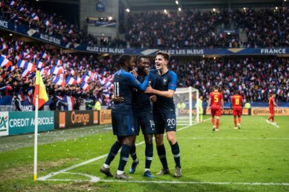 Trois joueurs de l'équipe de France en maillot bleu se congratulent près du poteau de corner, devant des tribunes pleines